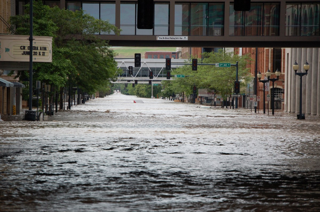Überschwemmte Stadtstraße mit Wasser, das die Straße, die Infrastruktur und Gebäude bedeckt, einschließlich einer Brücke im Hintergrund.