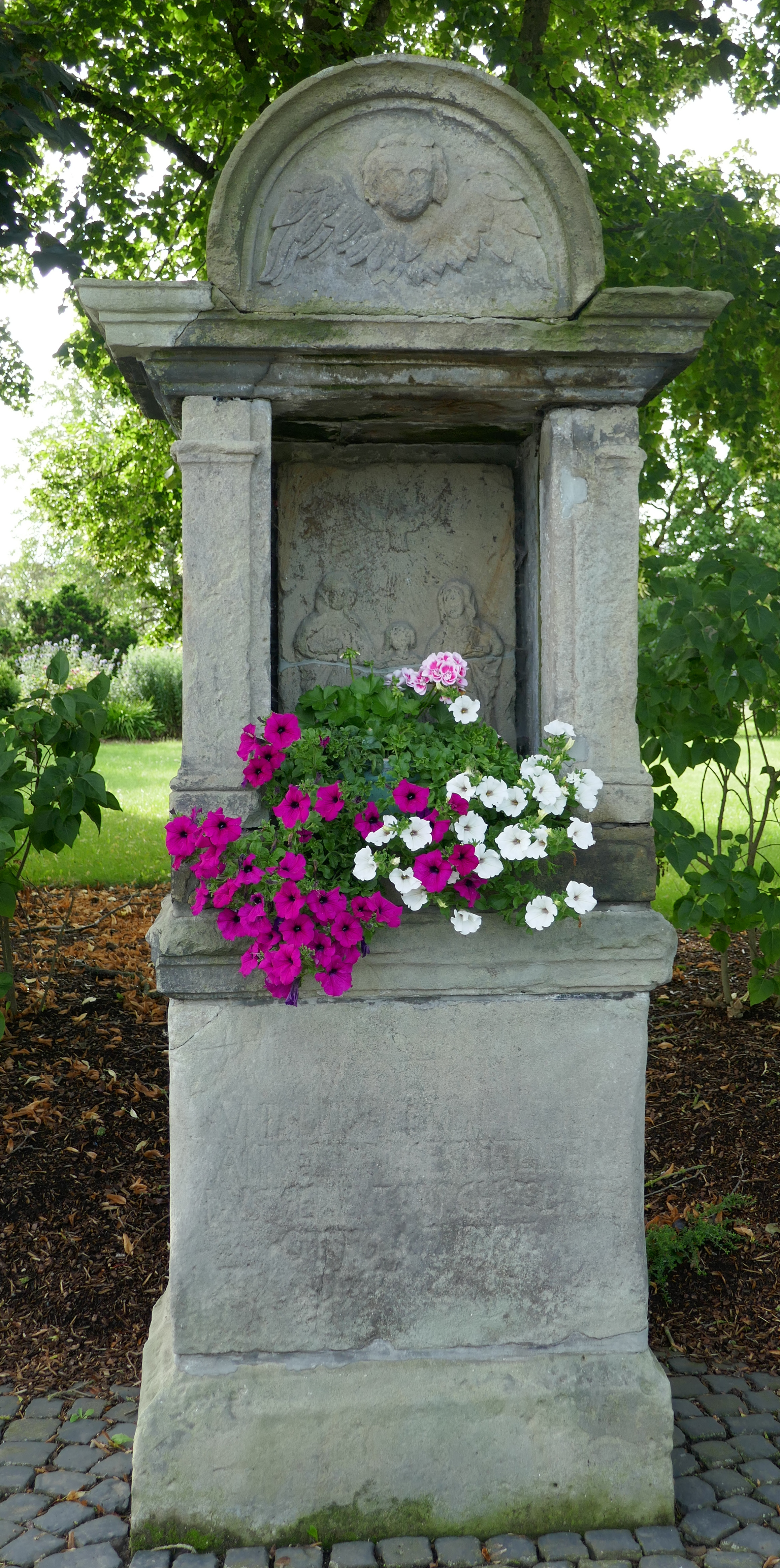 Ein steinerner Monument mit einem Blumentopf voller Blumen darauf, umgeben von Bäumen und einem klaren blauen Himmel.