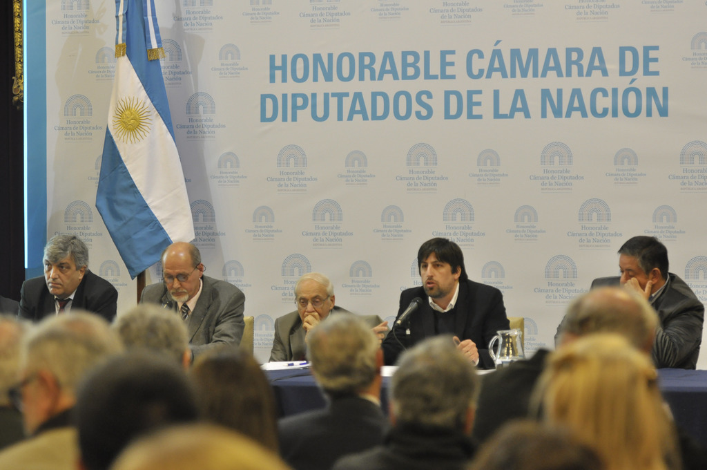 Eine Gruppe von Menschen sitzt an einem Tisch mit einem Krug, Papieren und einem Stift, während eine Person in der Mitte in ein Mikrofon spricht, mit einer weißen und blauen Flagge und einem Hintergrund, auf dem 'ehrenwerte kamara de' steht.