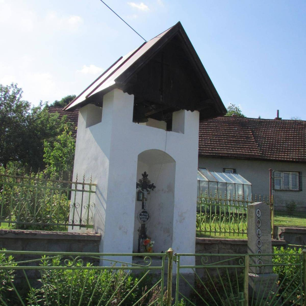 Kleines weißes Kirchengebäude mit kreuzbekröntem Dach, umgeben von einem Metallzaun, zwischen Pflanzen und Gras in der Nähe von Häusern unter einem bewölkten Himmel.