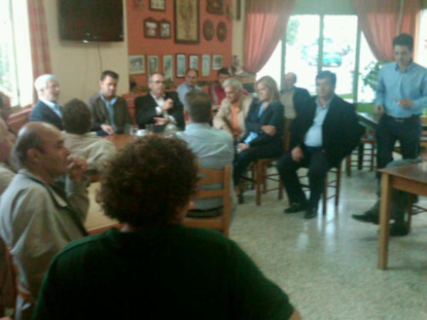 Eine Gruppe von Menschen sitzt in einem Raum mit einer Person in einem blauen Hemd, einem Fernseher, Gemälden an der Wand, einem Glasfenster mit Fahrzeugen und Bäumen und Vorhängen.