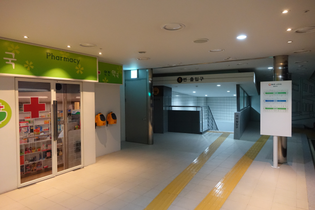 Der Eingang einer Apotheke mit einem "Apotheke"-Schild links, einer Tafel mit Text rechts und beleuchteten Lampen auf dem Dach.