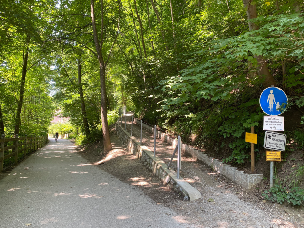 Eine Person fährt mit dem Fahrrad auf einem asphaltierten Weg durch den Wald, mit Bäumen und einem Holzzaun auf der linken Seite und Schildern mit Text und Bildern auf der rechten Seite.