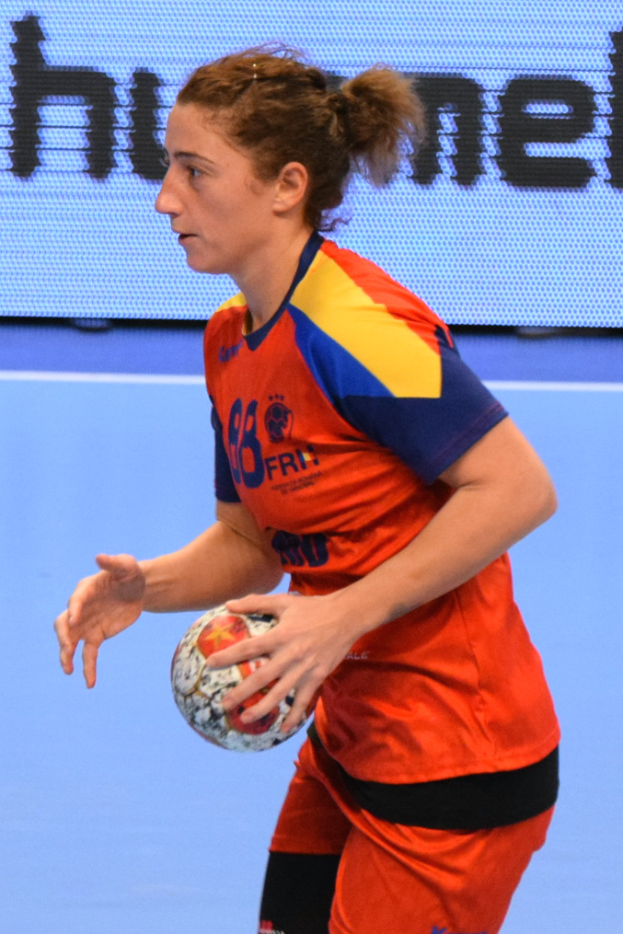 Eine Frau in einer orange-blauen Uniform hält einen Fußball, im Hintergrund ist ein Bildschirm mit Text zu sehen.