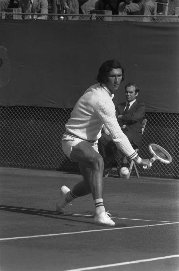Ein professioneller Tennisspieler in Sportschuhen schlägt mit einem Schläger nach einem Ball während eines Matches, mit Maschendrahtzaun und Zuschauern auf Stühlen im Hintergrund.