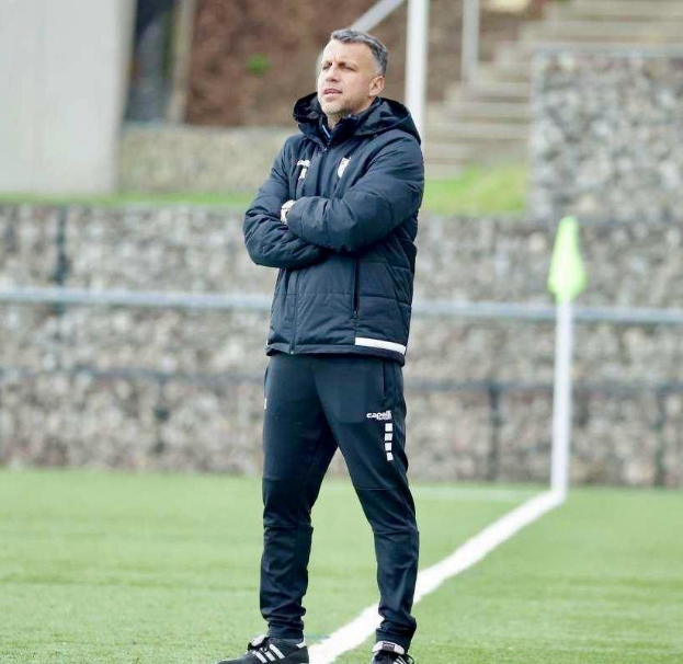 Ein Mann in schwarzer Jacke und Hose steht auf einem Fußballfeld mit verschränkten Armen, als wäre er ein Trainer während einer Trainingseinheit.