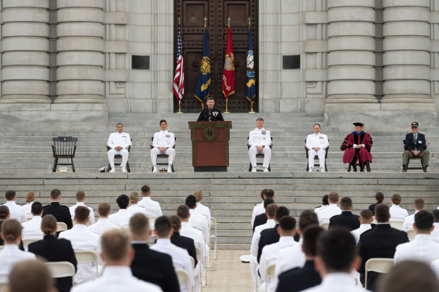 Gruppe von Menschen, die auf Stühlen vor einem Gebäude mit Säulen und einer Tür sitzen, mit einem Podium und einem Mikrofon in der Mitte, Fahnen im Hintergrund und einem leeren Stuhl links.