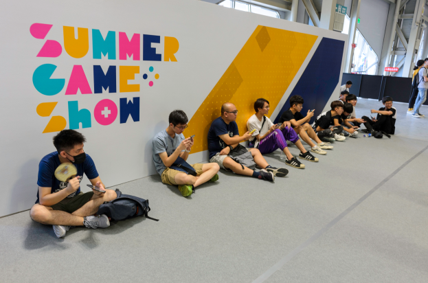 Eine Gruppe von Menschen sitzt vor einer Wand mit Text auf dem Boden, einige halten Mobiltelefone in der Hand, mit verstreuten Taschen und einigen Menschen im Hintergrund, beleuchtet von Deckenlampen.
