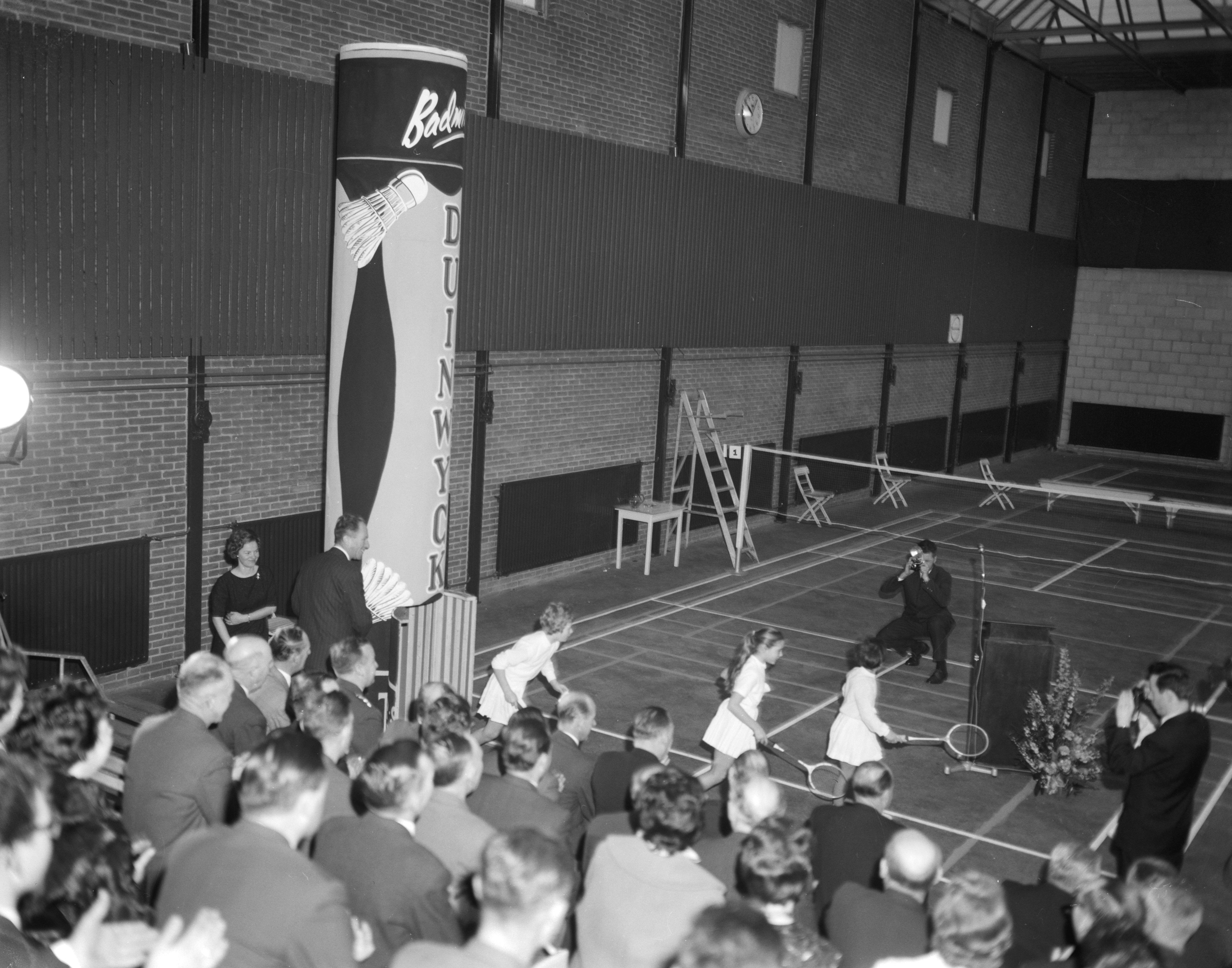 Ein Schwarz-Weiß-Foto einer Gruppe von Menschen, die Tennis in einer Turnhalle spielen, einige stehen und einige sitzen auf Stühlen mit Schlägern, mit einer Uhr an der Wand im Hintergrund.