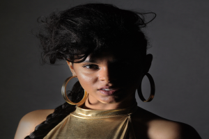 Eine Frau in einem goldenen Kleid und großen Kreolen steht selbstbewusst vor einer Wand, ihr Haar ist zu einem sleeken Bob gestylt und sie schaut direkt in die Kamera mit einem entschlossenen Ausdruck.