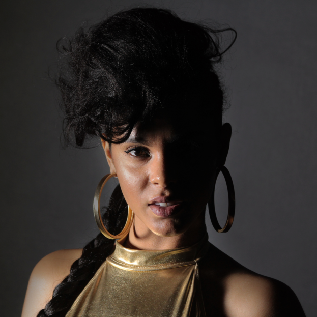 Eine Frau in einem goldenen Kleid und großen Kreolen steht selbstbewusst vor einer Wand, ihr Haar ist zu einem sleeken Bob gestylt und sie schaut direkt in die Kamera mit einem entschlossenen Ausdruck.