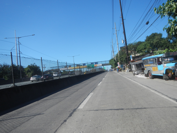 Straßenkreuzung mit fahrenden Autos, geparktem Bus, Strommasten mit Drähten, einem Zaun auf der linken Seite, Bäumen, Laternen, einer Brücke im Hintergrund und einem blauen Himmel.