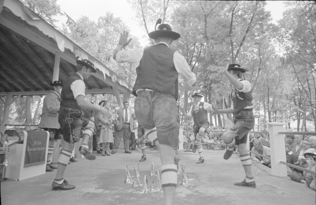 Eine Gruppe von Männern in traditioneller bayrischer Tracht tanzt auf einer Bühne vor einem gemischten Publikum aus sitzenden und stehenden Menschen, mit Trees und einer Hütte im Hintergrund, dargestellt in Schwarz-Weiß.