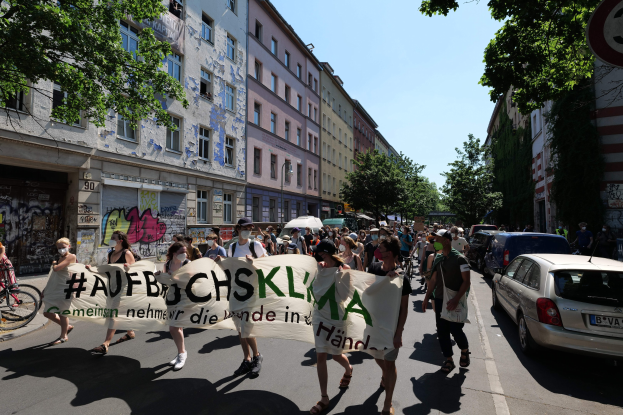 Eine Gruppe von Menschen marschiert mit einem Banner die Straße entlang, mit parkenden Autos auf der rechten Seite, einem Fahrrad auf der linken Seite und Gebäuden, Bäumen und einem Schild im Hintergrund.