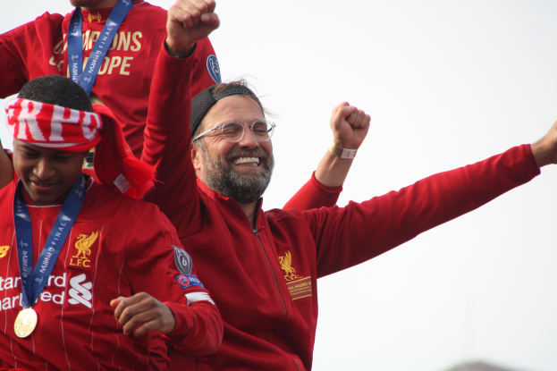 Liverpool-Trainer Jürgen Klopp feiert den Sieg in der UEFA Champions League mit seinen Spielern, während zwei medalletragende Personen in der Nähe sind und der Himmel im Hintergrund zu sehen ist.