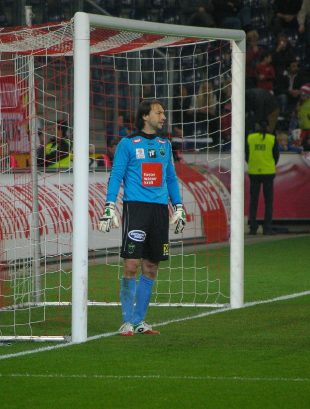 Ein Fußballspieler in einem blauen T-Shirt, schwarzen Shorts, Handschuhen und Schuhen steht vor einem Tor auf einem Rasenfeld, mit Zuschauern auf Stühlen und Stehplätzen im Hintergrund und ein paar Schildern mit Text.