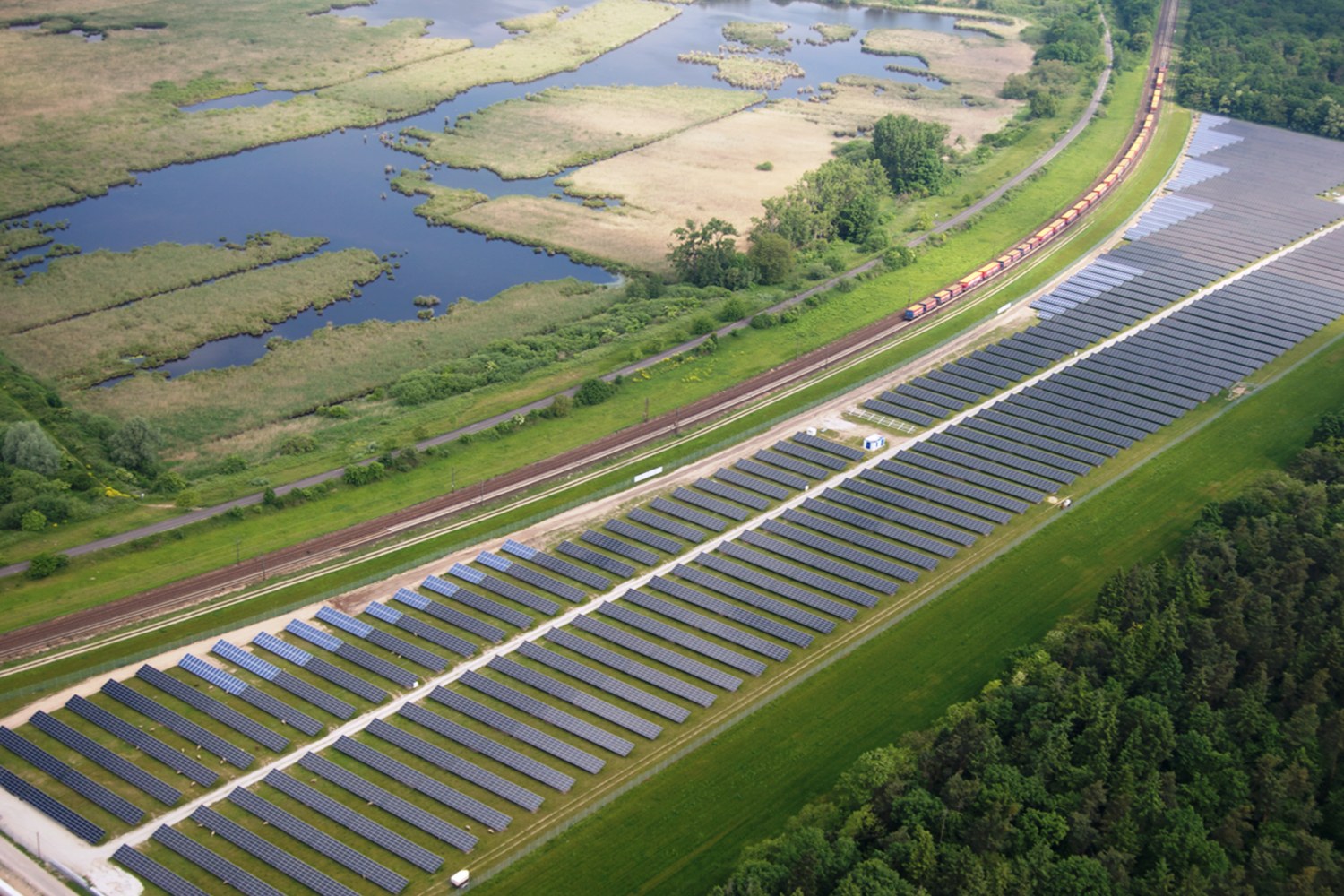 Luftbild einer Solarstromanlage mit Panelen in einem Feld, umgeben von Bäumen, Gras, Pflanzen und Wasser, mit einem Zug auf einer nahen Bahnstrecke.