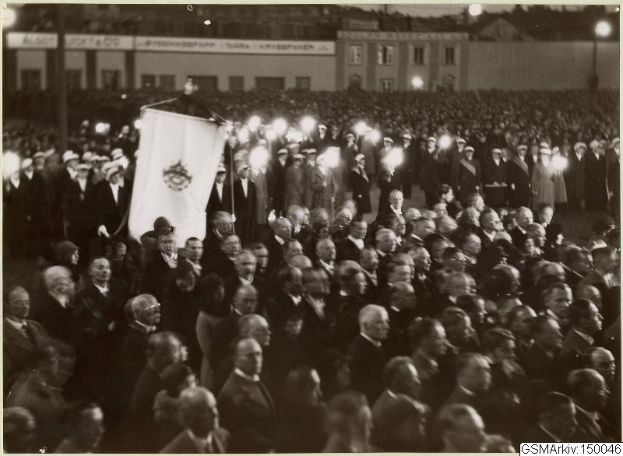 Schwarzes und weißes Foto einer großen Menge, die vor einem Gebäude steht, mit einer Person, die eine Flagge hält, und Text, der unten "Gustav Klimts Rede an der Berliner Mauer" lautet.
