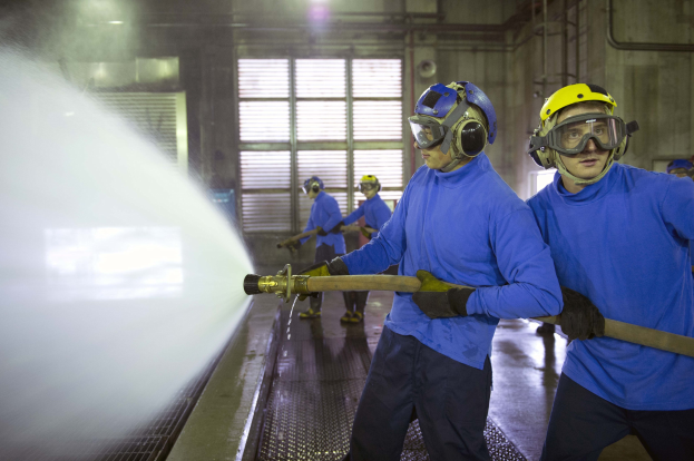 Männer in blauen Hemden und gelben Helmen arbeiten an Maschinen, einer sprüht Wasser auf den Boden, in einer industriellen Fabrikumgebung mit Rohren, Fenstern und Lampen.