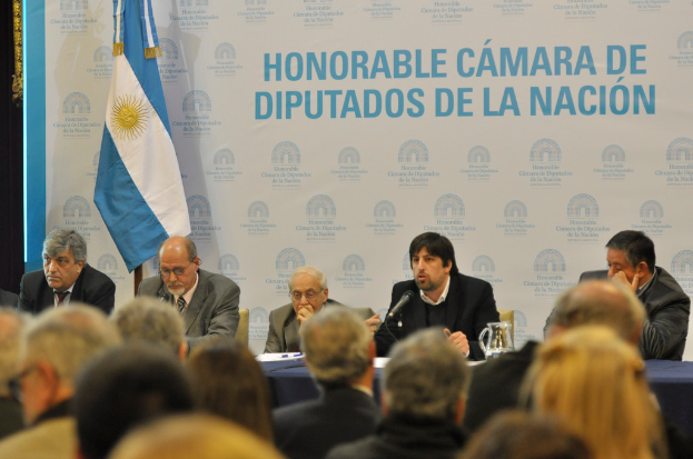 Eine Gruppe von Menschen sitzt in Reihen von Stühlen, mit einem Tisch vor der ersten Reihe, der einen Krug, Papiere und einen Stift enthält. Eine Person in der Mitte spricht in ein Mikrofon, mit einer weißen und blauen Flagge dahinter und einem weißen und blauen Hintergrund, auf dem "ehrenwerte kamara de" geschrieben steht.