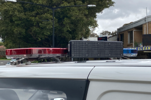 Ein Polizeiwagen mit eingeschaltetem Blaulicht steht am Straßenrand, umgeben von Gebäuden, Bäumen, Polen und Schildern, unter einem bewölkten Himmel, wahrscheinlich nach einer tödlichen Schulschießerei in Brisbane.