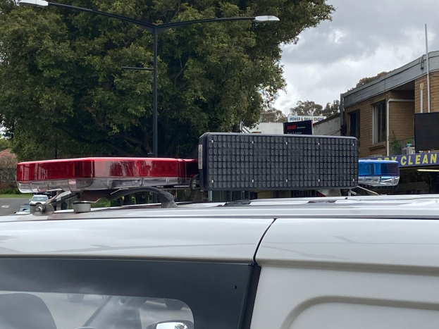 Ein Polizeiwagen mit eingeschaltetem Blaulicht steht am Straßenrand, umgeben von Gebäuden, Bäumen, Polen und Schildern, unter einem bewölkten Himmel, wahrscheinlich nach einer tödlichen Schulschießerei in Brisbane.