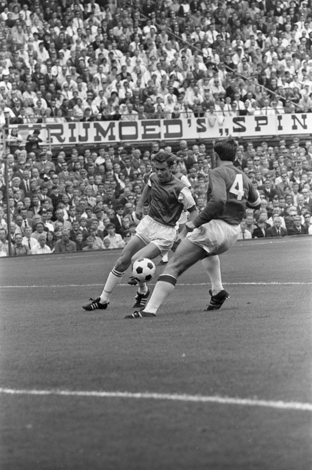 Schwarzes und weißes Foto von zwei Männern, die auf einem Feld Fußball spielen, mit Zuschauern im Hintergrund und einem Banner mit der Aufschrift "Bobby Charlton im Einsatz für Manchester United."