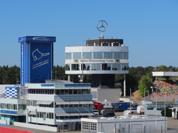 Ein Blick auf das Hauptquartier des Mercedes-AMG Petronas F1 Teams in Berlin, Deutschland, mit Gebäuden, Fahrzeugen auf der Straße, einem Zaun, Pfählen, Tafeln mit Text, Bäumen und einem Logo auf dem Gebäude, mit dem Himmel im Hintergrund.