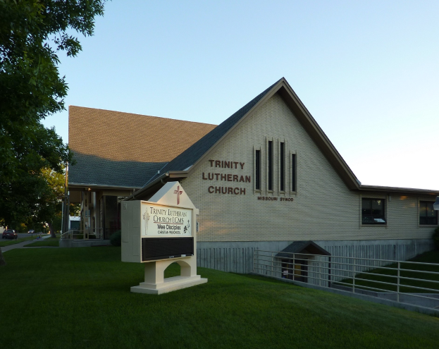 Außenansicht der Trinity Lutheran Church in St. Louis, Missouri, mit einem Schild, einem Metallzaun, Gras, einer Straßenlaterne, Bäumen, Fahrzeugen und einem bewölkten Himmel.