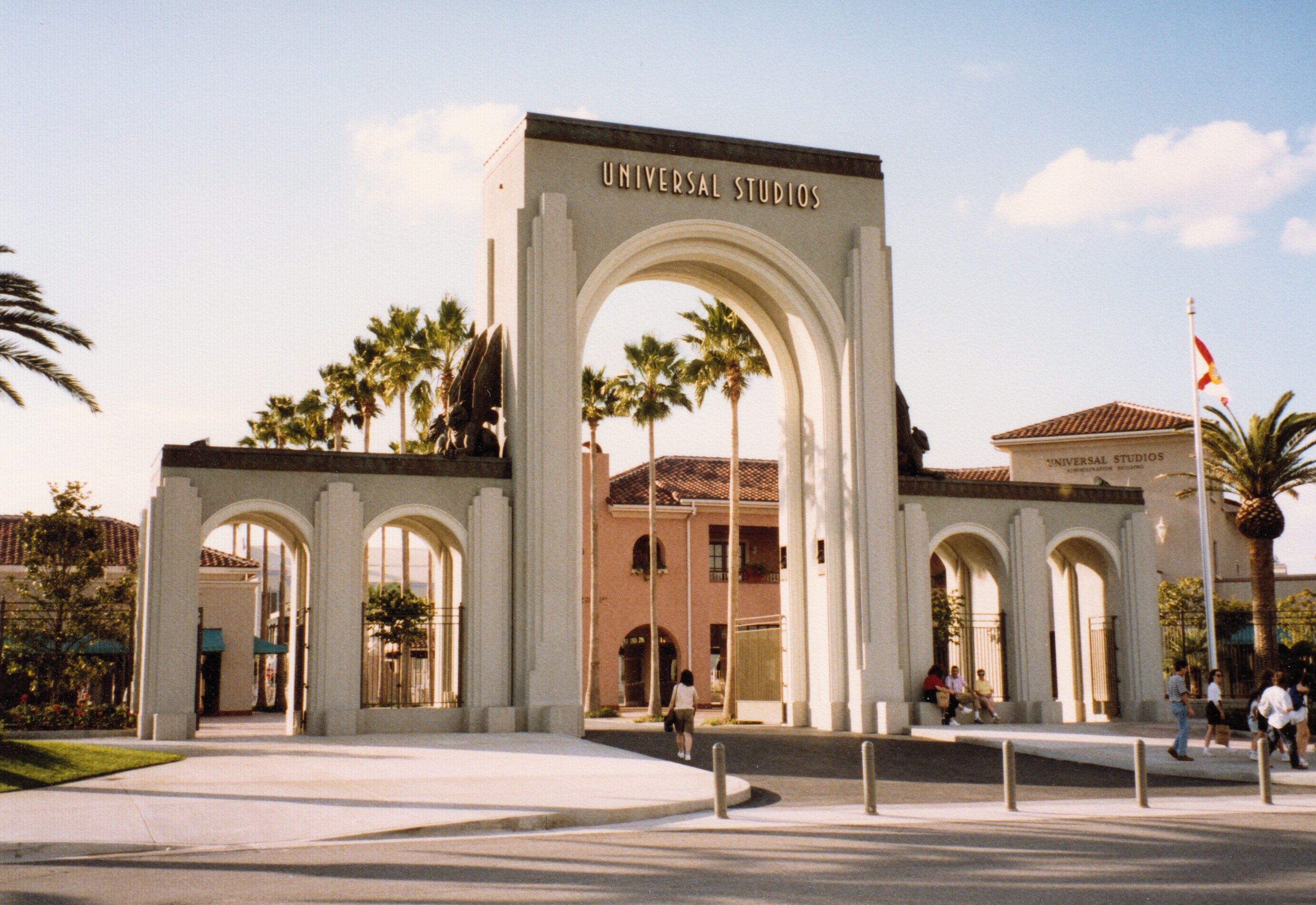 Eingang zu Universal Studios Hollywood mit einem Bogen mit Text, einer Statue, Menschen, Bäumen, Gebäuden, einem Fahnenmast und einem bewölkten Himmel.