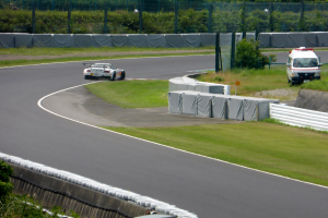 Ein Rennwagen fährt auf einer Rennstrecke mit einem Polizeiwagen im Hintergrund, umgeben von Gras, Pflanzen, Bäumen und Zäunen, mit einem Fahrzeug auf der rechten Seite und Text unten.