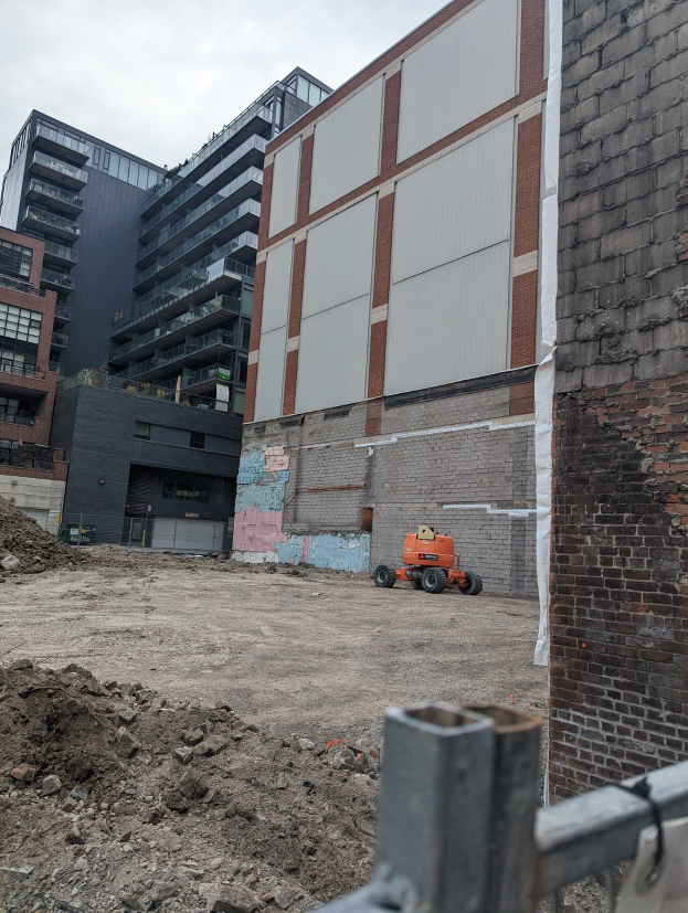 Baustelle mit einem großen Gebäude unter Abriss im Hintergrund, einem Fahrzeug auf dem Boden, einem Metallzaun und einem bewölkten Himmel.