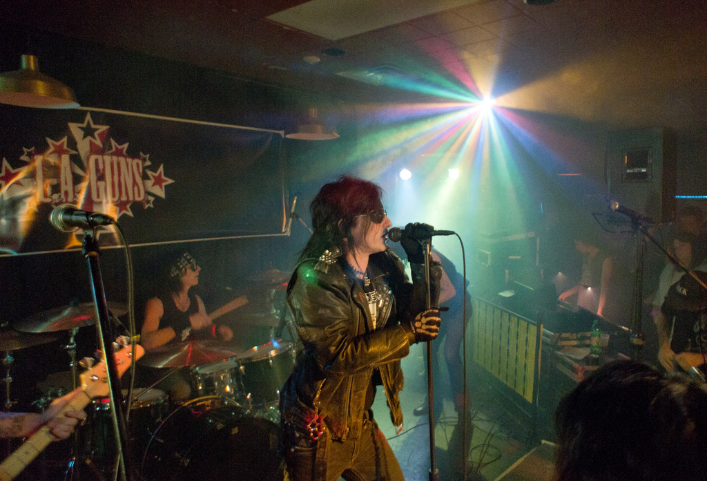 Eine Frau in einer schwarzen Jacke singt in ein Mikrofon auf der Bühne, mit einem Mann in Sonnenbrille und Stirnbinde, der ein Instrument spielt; ein "LA Guns"-Plakat und Diskoleuchten sind im Hintergrund zu sehen.