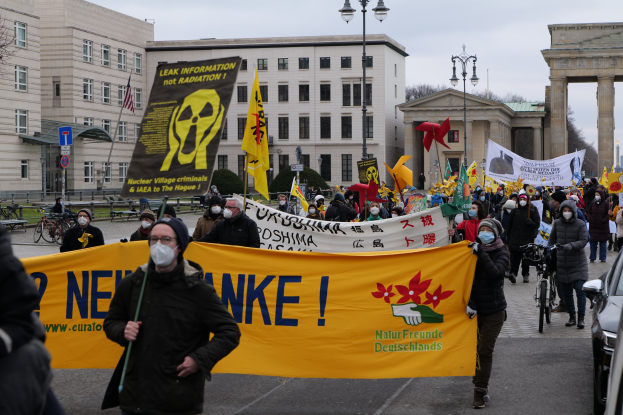 Große Gruppe von Menschen bei einer Straßendemonstration gegen Atomkraft in Deutschland, mit Transparenten und Fahnen, Fahrzeugen auf der rechten Seite, Gebäuden und Bäumen im Hintergrund und einem Tor weiter unten auf der Straße.
