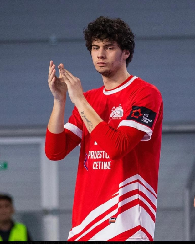 Ein Mann in einem roten und weißen T-Shirt klatscht in den Händen in der Vordergrund mit einer anderen Person und einer Wand im Hintergrund, mit Text, der unten "Copenhagen Futsal Club - Foto 1" liest.