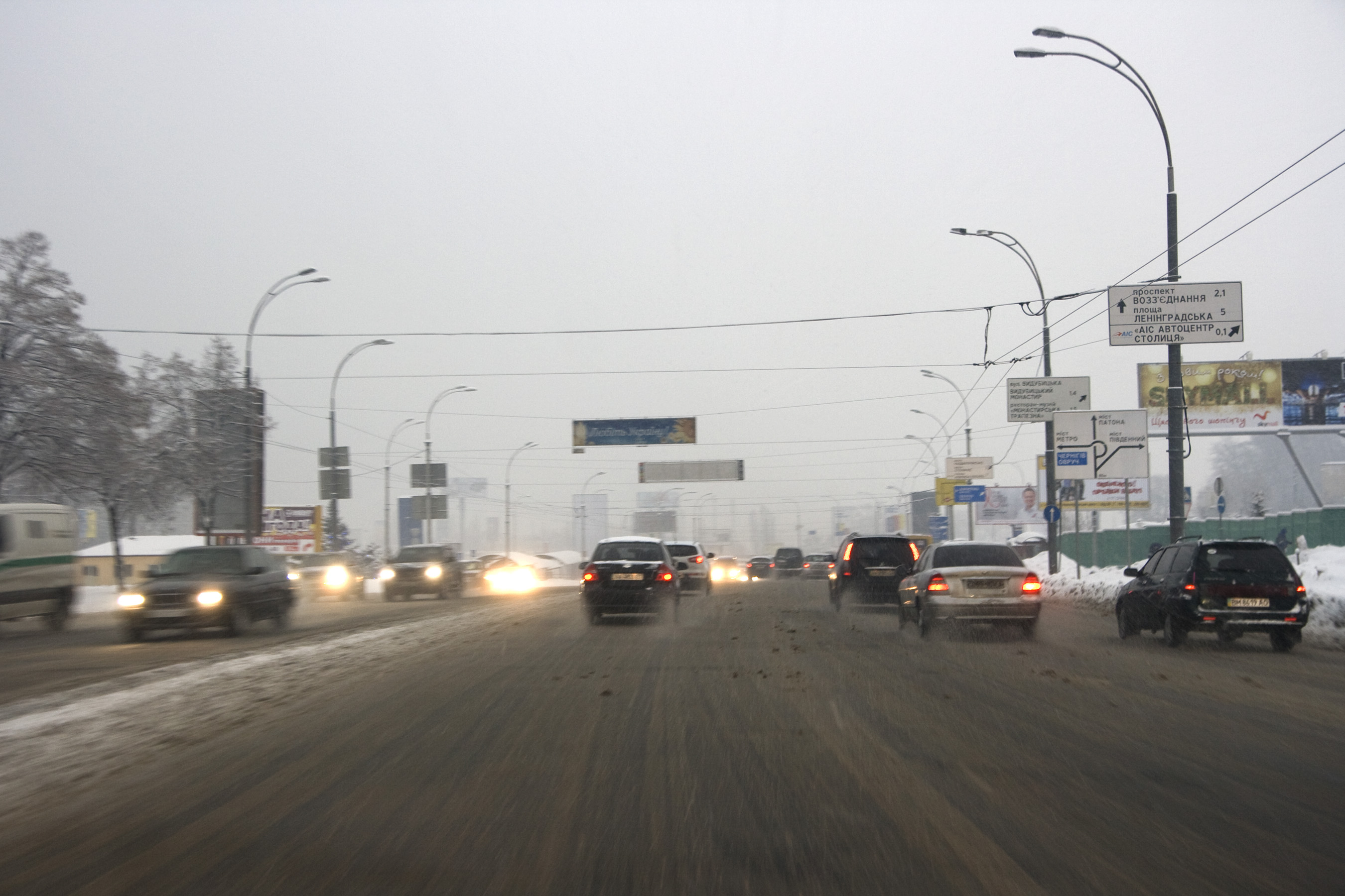 Eine belebte Stadtstraße mit Fahrzeugen auf einer schneebedeckten Straße, gesäumt von Laternen, Texttafeln, Bäumen und Gebäuden unter einem bewölkten Himmel.