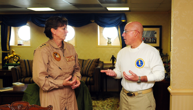 Ein Mann und eine Frau unterhalten sich in einem Wohnzimmer mit Möbeln, Bücherregalen und einem Foto auf dem Wand, mit Fenstern und Deckenlampen im Hintergrund.