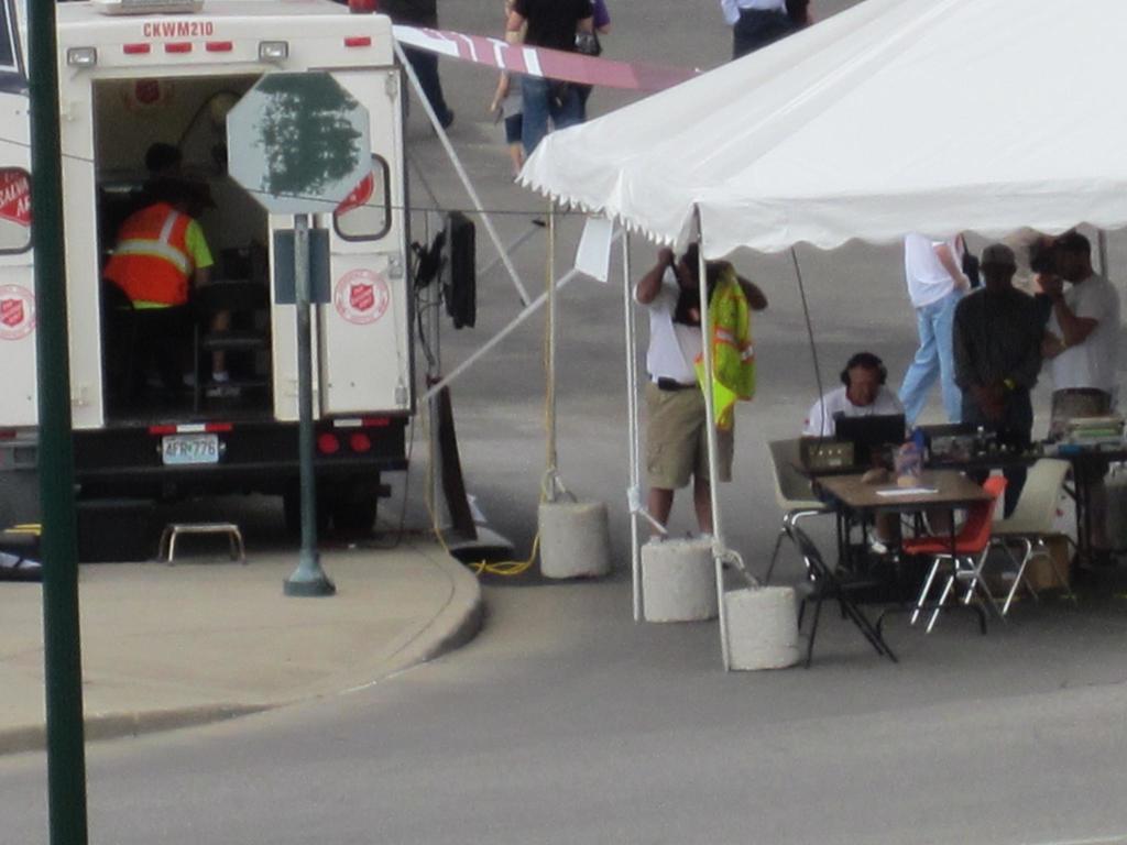 Ein Krankenwagen steht auf der linken Seite einer Straße, während auf der rechten Seite ein Zelt eine Gruppe von Menschen schützt, mit einem Tisch und einem Stuhl in der Nähe.