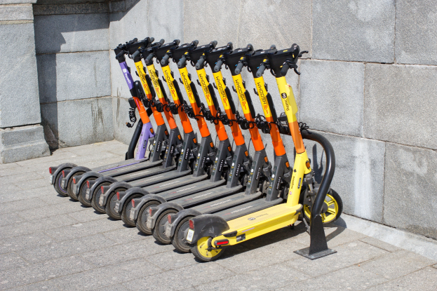 Eine Reihe von Elektro-Scootern ordentlich in einer Reihe auf einem Gehweg geparkt, mit einer Wand im Hintergrund.