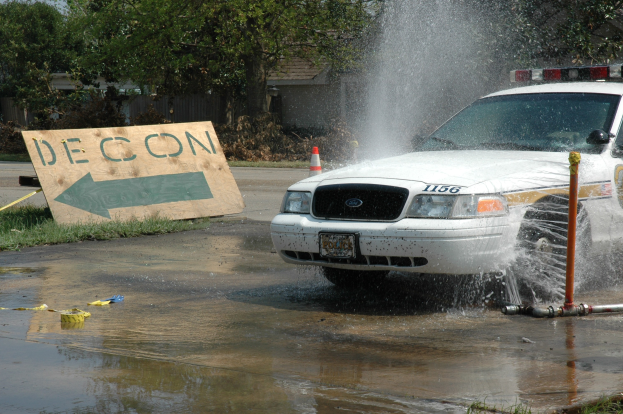 Ein Streifenwagen fährt durch eine Pfütze, mit einem Schild, einer Verkehrskegel und anderen Gegenständen auf dem Boden, vor einem Hintergrund aus Bäumen, Gebäuden und einem klaren blauen Himmel.