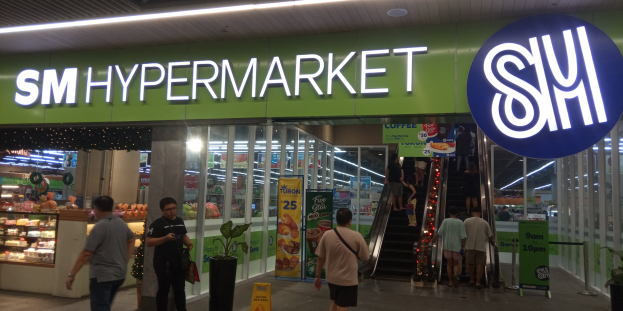 Eine Gruppe von Menschen, darunter Männer und Frauen, geht vor einem hell erleuchteten Geschäft mit einem Schild, das 'SM Hypermarket' lautet, vorbei, mit sichtbaren Tafeln, Topfpflanzen und anderen Gegenständen im Inneren.
