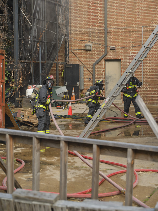 Feuerwehrleute in Helmen arbeiten mit Handspritzen, um ein Gebäude Feuer zu löschen, mit einem Metallzaun, verstreuten Rohren, einem Behälter, einem Verkehrskegel und Hintergrundelementen wie einem Baum und Himmel.