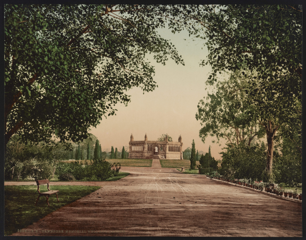 Ein altes Foto eines Parks mit einer Bank, umgeben von grünem Gras, Pflanzen und Bäumen, mit einem Gebäude mit Säulen und Bögen im Hintergrund und Text am unteren Rand des Bildes.