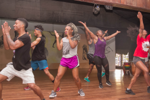 Eine Gruppe von Menschen, die in einem Tanzstudio tanzen, mit einem Gemälde an der Wand, einem Glasfenster auf der rechten Seite und Deckenleuchten darüber, was auf eine Zumba-Stunde hinweist.