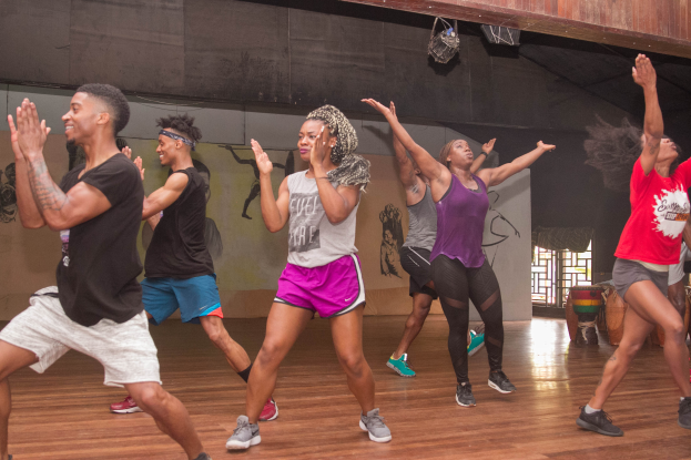 Eine Gruppe von Menschen, die in einem Tanzstudio tanzen, mit einem Gemälde an der Wand, einem Glasfenster auf der rechten Seite und Deckenleuchten darüber, was auf eine Zumba-Stunde hinweist.