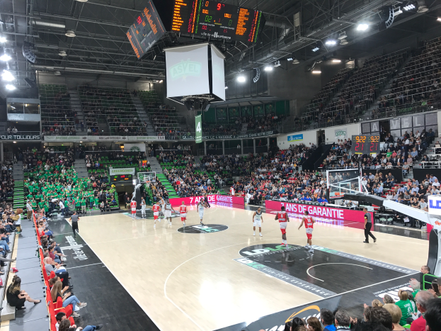 Ein Basketballspiel in einer grossen Arena mit Zuschauern auf den Rängen, Anzeigetafeln mit "Asvel vs Galatasaray" und einer Decke mit Lampen und einem Bildschirm.