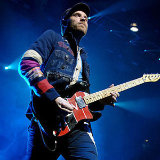 Ein Mann mit Mütze spielt Gitarre auf der Bühne eines Konzerts, beleuchtet von hellen Scheinwerfern, mit einem freudigen und aufgeregten Gesichtsausdruck.