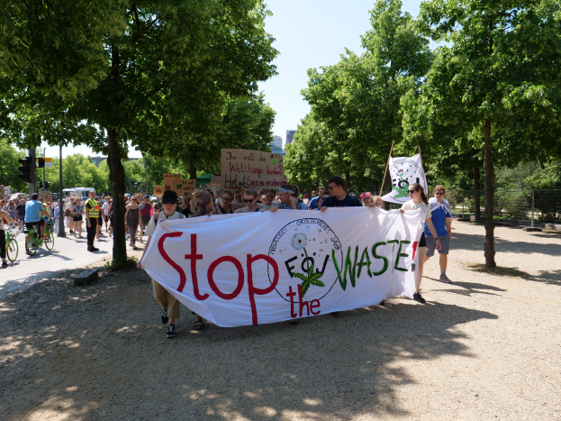 Eine Gruppe von Menschen marschiert eine von Bäumen gesäumte Straße entlang, hält eine "Stop the Waste"-Tafel und verschiedene Plakate, während einige Fahrräder fahren, Gebäude im Hintergrund unter einem klaren blauen Himmel.