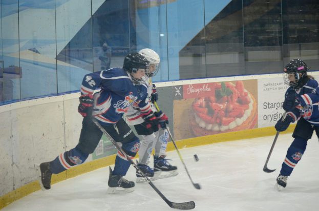 Gruppe junger Menschen, die Eis hockey auf einer Indoor-Eisfläche spielen, mit Helmen, Sportkleidung und Hockey-Schlägern, sowie einem Plakat im Hintergrund an einer Glaswand.