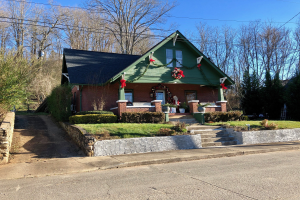 Ein grünes Haus mit einem Kranz an der Vorderseite, umgeben von Pflanzen, Gras, Treppen, einer Wand, Bäumen und Drähten, unter einem bewölkten Himmel, gelegen im Herzen der Stadt und zur Miete verfügbar.